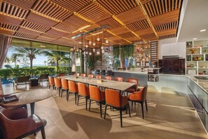 A modern dining area with a long table and orange chairs, a wooden ceiling with hanging lights, and a view of palm trees outside. - 8BR Beachfront Eden: Luxury Events & Getaways (Da Nang)