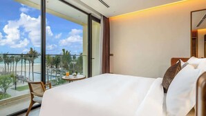 A hotel room with a large bed, a small table with a drink, a chair, and a balcony with a view of the beach and palm trees.