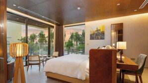 A modern hotel room with a large bed, a desk, and a chair. There is a large window offering a view of palm trees and a sunset.
