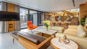 A modern living room with a sofa, coffee table, and orange chair. There is a dining area with a staircase and a TV mounted on the wall.