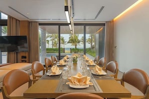 Une salle à manger avec une longue table dressée pour le repas, de grandes fenêtres offrant une vue sur les palmiers et l’océan, et un téléviseur à écran plat fixé au mur.