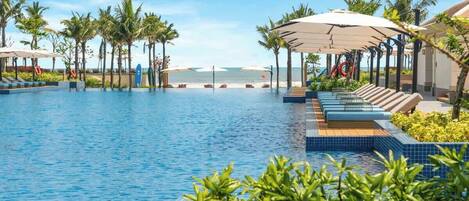 A swimming pool with lounge chairs and umbrellas, surrounded by palm trees and a view of the ocean.