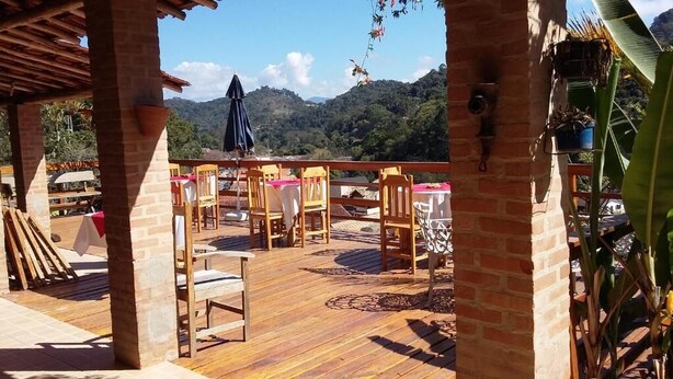 An outdoor patio with wooden tables and chairs, a brick wall, and a view of green hills. - Pousada Pinhalense (Santo Antonio do Pinhal)
