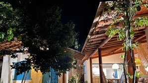 A well-lit outdoor area with a wooden pergola, a dining area with chairs and a table, and a building with blue shutters. - Trancoso Biriba guest house (Porto Seguro)