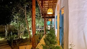 A covered outdoor area with a wooden ceiling and a white wall with a blue door. There are plants and a pathway leading to a building. - Trancoso Biriba guest house (Porto Seguro)