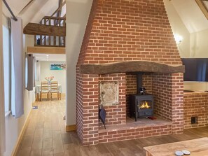 A brick fireplace with a wood-burning stove, a dining area with a table and chairs, and a lofted wooden staircase. - The Dairy at Spratts Green Farm (Aylsham)