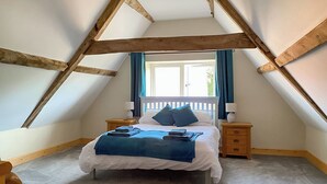 Ein Schlafzimmer mit schrägen Wänden, einem Bett mit blauer Bettwäsche, zwei Nachttischen mit Lampen und einem Fenster mit Vorhängen.