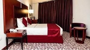 Habitación de hotel con una cama grande, una mesita de noche con lámpara, una silla y una alfombra estampada.