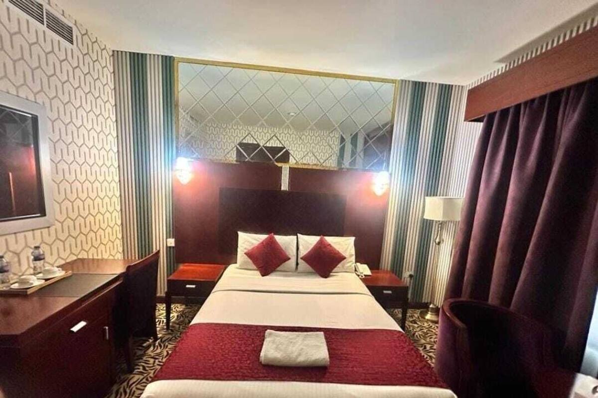 A hotel room with a bed, a desk, a chair, a nightstand, a lamp, and a mirror.