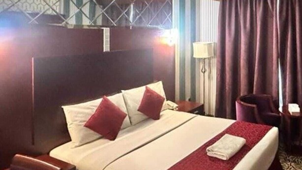 Habitación de hotel con una cama grande, cabecera de madera, una manta roja y alfombra estampada.
