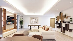 Een moderne woonkamer met een bank, salontafel en een pooltafel. Links een televisie en rechts een boekenkast.