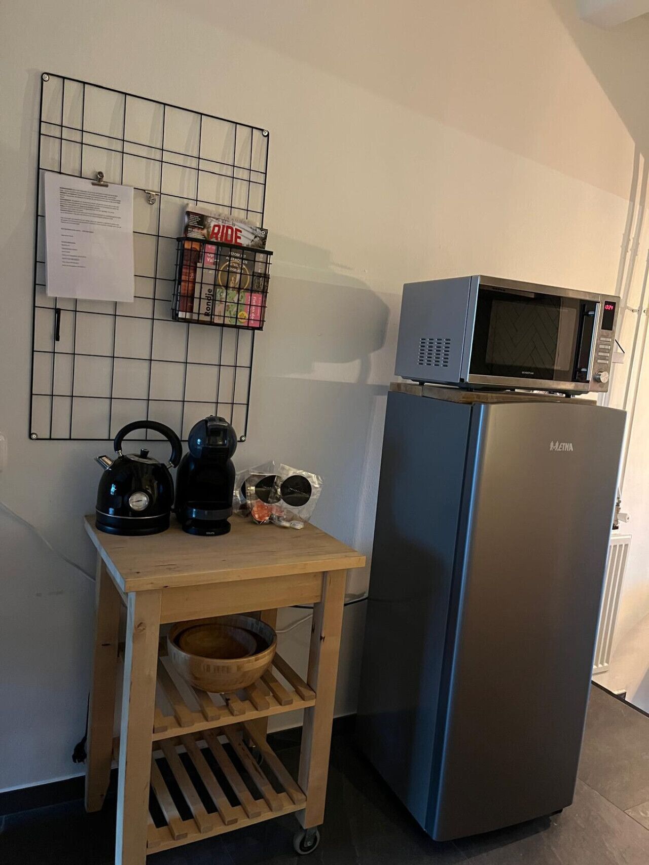 A kitchen with a microwave on top of a refrigerator, a kettle, a coffee maker, and a basket of bread on a wooden cart.
