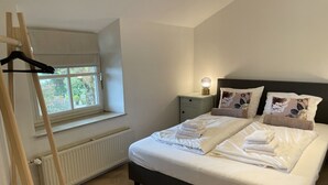 Quarto com cama, mesa de cabeceira, abajur e janela com vista para o verde.