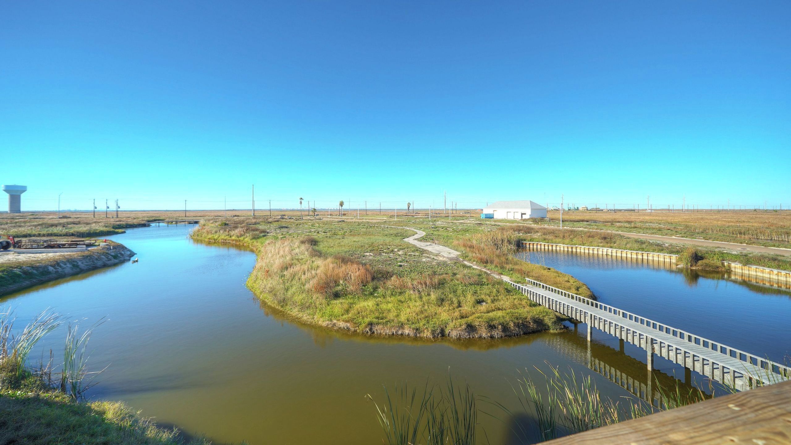 A river with a wooden bridge, a white building, and a water tower in the background.