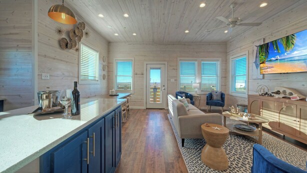 A spacious living area with a kitchen, wooden floors, and a large TV screen displaying a beach scene.
