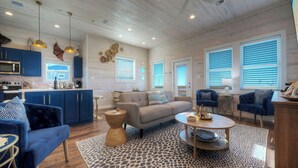 A spacious living area with a sofa, coffee table, and blue armchairs. A kitchen with blue cabinets and a microwave is visible.