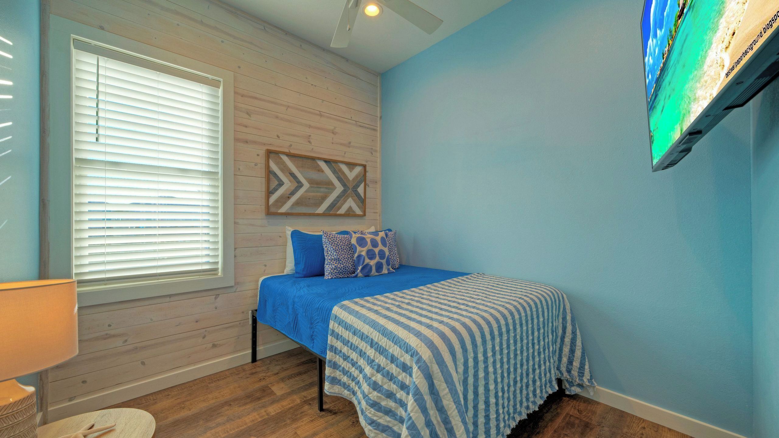 Une chambre à coucher avec un lit, une fenêtre avec des stores, un téléviseur fixé au mur et un plancher en bois.
