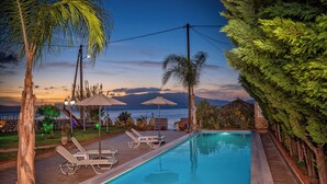 A swimming pool with lounge chairs and umbrellas, surrounded by trees and a view of the ocean at sunset.