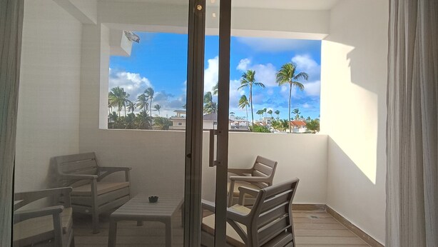 A balcony with a view of palm trees and buildings, wooden furniture, and a glass door.