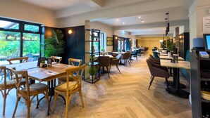 Restaurante moderno com mesas e cadeiras de madeira, uma estante preta e grandes janelas com vista para o verde.