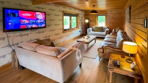 Interior acolhedor de uma cabana de madeira com uma televisão de tela plana grande fixada na parede, uma área de estar confortável com sofás e uma mesa de centro, e pisos de madeira.
