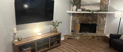 Ein Wohnzimmer mit flachbildfernseher an der Wand, einem Kamin aus Stein mit Kaminfeuerholzablage und einer Holzunterhaltungsecke.