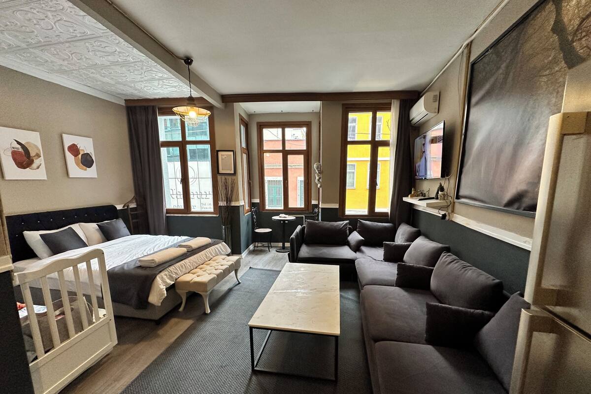 Ein Hotelzimmer mit Bett, Sofa, kleinem Tisch und Babybett. Es gibt Fenster mit Vorhängen, eine Wandklimaanlage und ein Bild an der Wand.