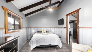 A bedroom with a bed, wooden ceiling beams, a ceiling fan, a dresser, a TV, and a door leading to another room. - Unique rural cabin, 5 minutes from beach (Valla)