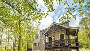 Une maison à deux étages dotée d’une grande terrasse et d’un porche abrité, entourée d’arbres et d’une pelouse.