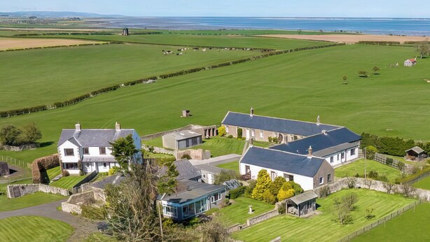 A large estate with multiple buildings, including a main house and several outbuildings, surrounded by expansive green fields and a coastal view. - Six the Square - UK48869 (Belford)