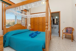 A four-poster bed with a blue bedspread, a wooden nightstand with a lamp, and a chair with a patterned cushion.