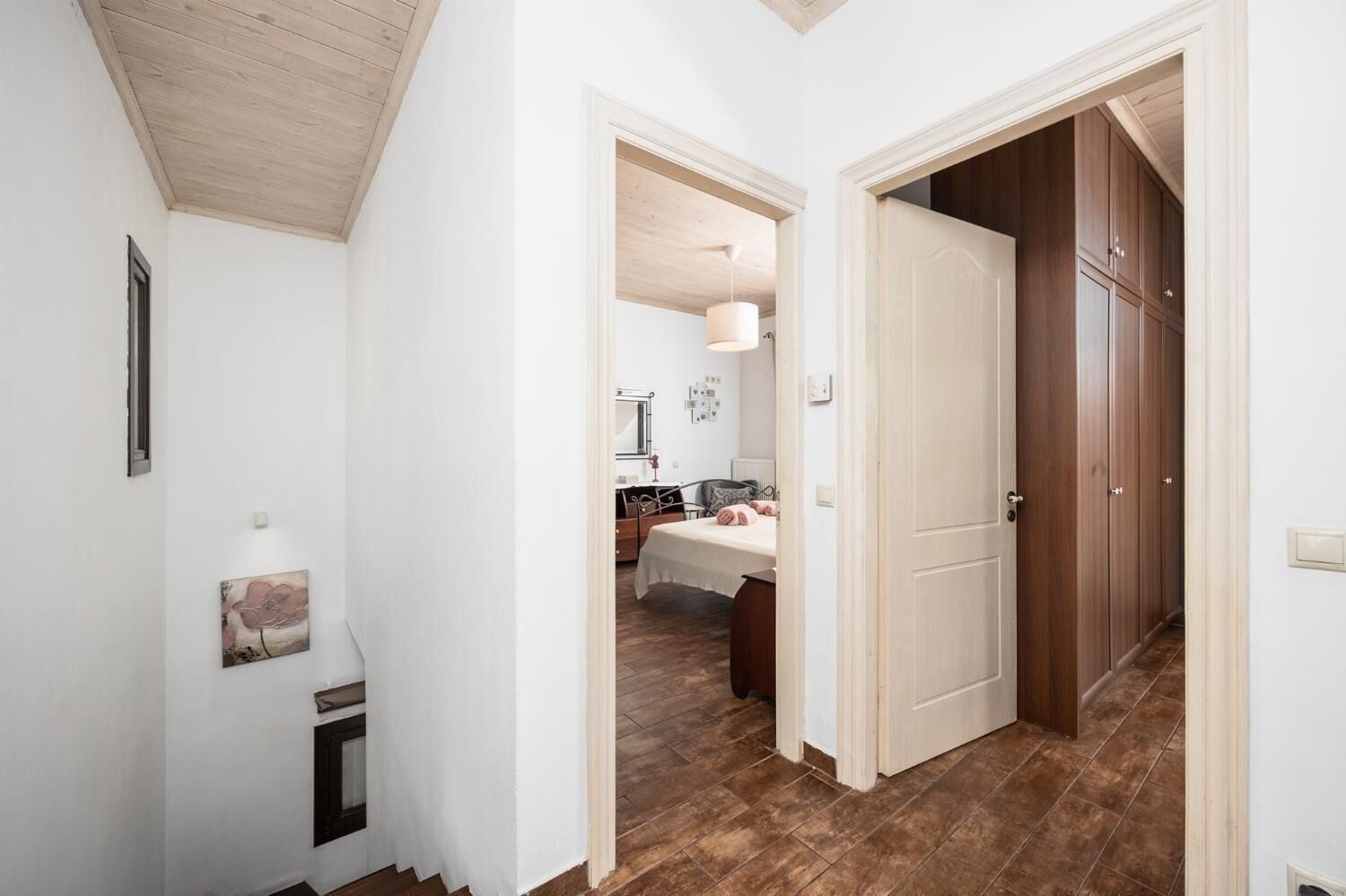 Un couloir avec une porte en bois qui donne sur une chambre à coucher, où se trouvent un lit, une table de chevet et un miroir.