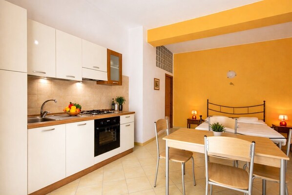 A compact kitchen with modern appliances, a dining area with a table and chairs, and a bedroom with a bed and bedside lamps.