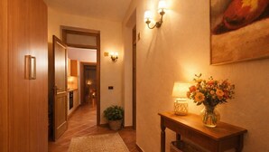 Ein Flur mit Holztüren, gefliestem Boden, einer Wandlampe, einem Tisch mit Lampe und Blumen sowie einem gerahmten Bild an der Wand.