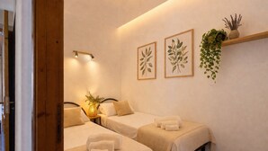 Habitación de hotel con dos camas, una mesita de noche con lámpara, estante en la pared con plantas e impresiones botánicas enmarcadas.