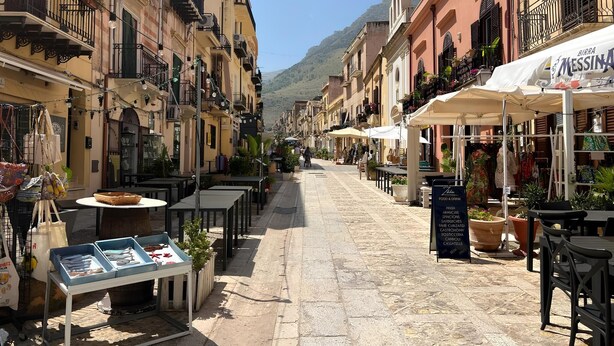 A narrow street lined with buildings, outdoor seating, and a shop with a sign. - Visitsicilia 6 Le Magnole 6 (Castellammare del Golfo)