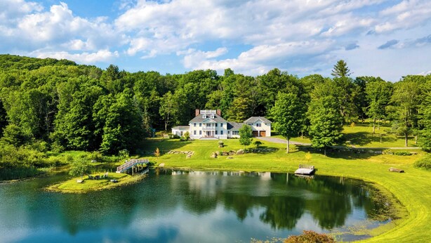 Une grande maison avec un étang devant, entourée d’une végétation luxuriante et d’une colline boisée.
