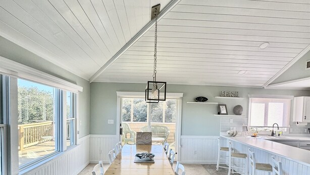 A spacious dining area with a long wooden table and white chairs, a kitchen with a white countertop and bar stools, and large windows offering a view of the outdoors. - 4x4 Carova, WaterFront, Dock, Wild Horses, Hot Tub (Corolla)