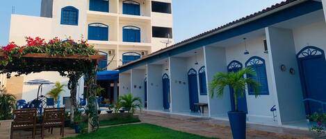 A courtyard with a white building featuring blue doors and windows, surrounded by greenery and outdoor seating.