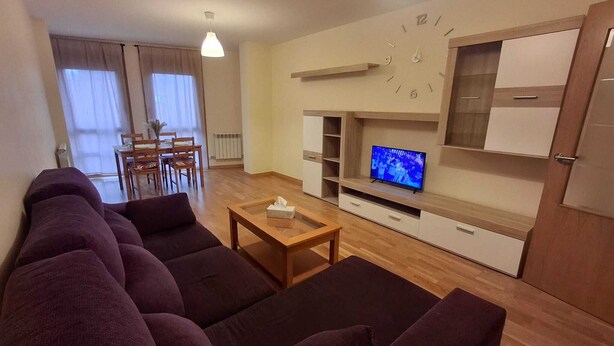 Ein modernes Wohnzimmer mit einer Sofagarnitur, einem Couchtisch, einem Fernseher auf einer Holzablage und einem Essbereich im Hintergrund.
