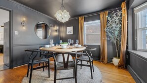 A dining area with a round table set for six, surrounded by dark chairs. A large mirror hangs on the wall, and a chandelier is visible above the table. - Park 4 Cars < > A Stunning Old Town Xcape (Alexandria)
