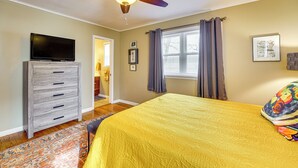 Un dormitorio con una cama, un ventilador de techo, un televisor sobre un tocador y una ventana con cortinas.