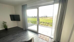 Una sala moderna con piso de madera oscura, un sofá gris, un televisor de pantalla plana montado en la pared y un gran ventanal corredizo que da a un área exterior con césped.