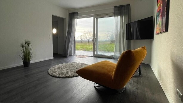Una sala de estar moderna con piso de madera oscura, un sillón color mostaza, un televisor de pantalla plana montado en la pared y un amplio ventanal con cortinas.