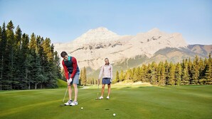 Golfkenttä, jolla kaksi henkilöä seisoo viheriöllä. Taustalla näkyy vuoristo ja vasemmalla puolella metsäinen alue.
