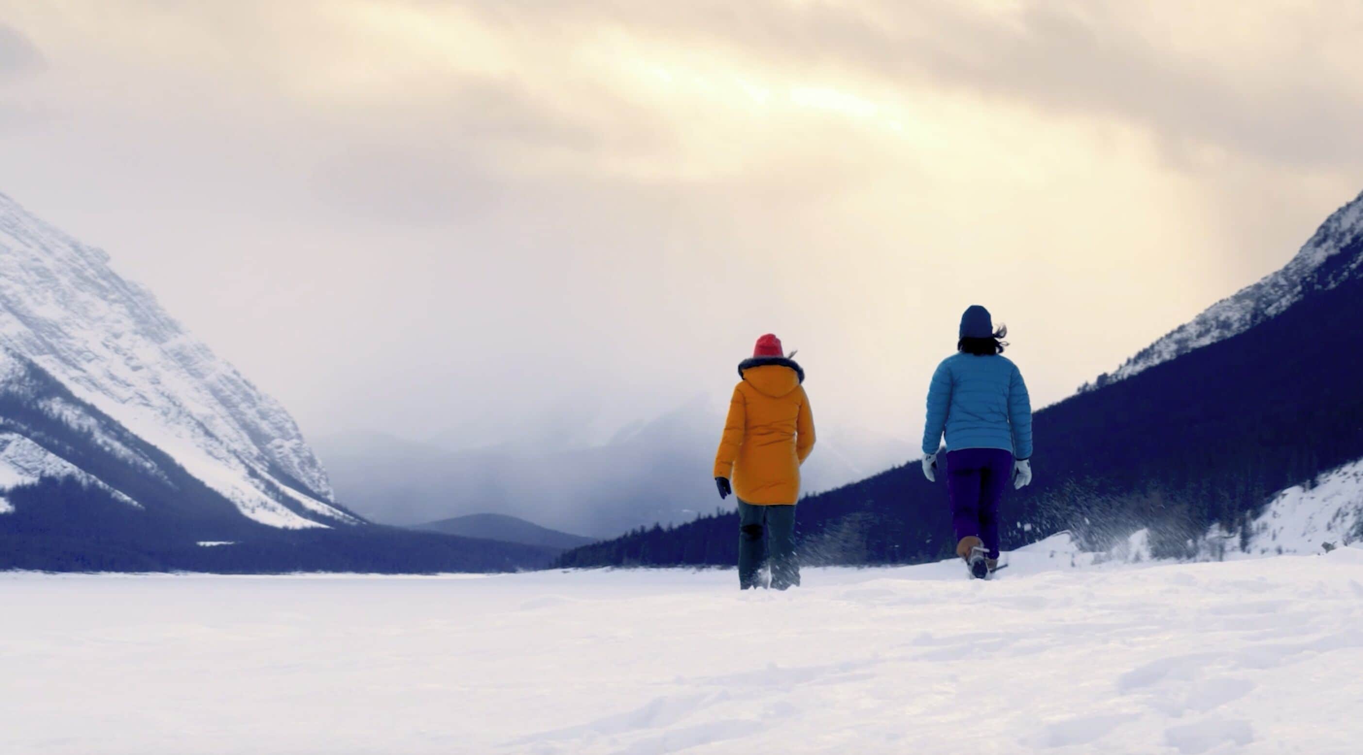 Two people walking on a snowy path with mountains in the background.