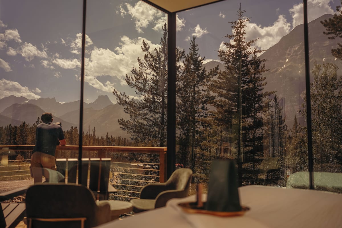Ein Zimmer mit großem Fenster, das auf eine gebirgige Landschaft blickt. Im Vordergrund befinden sich mehrere Stühle und ein Tisch.