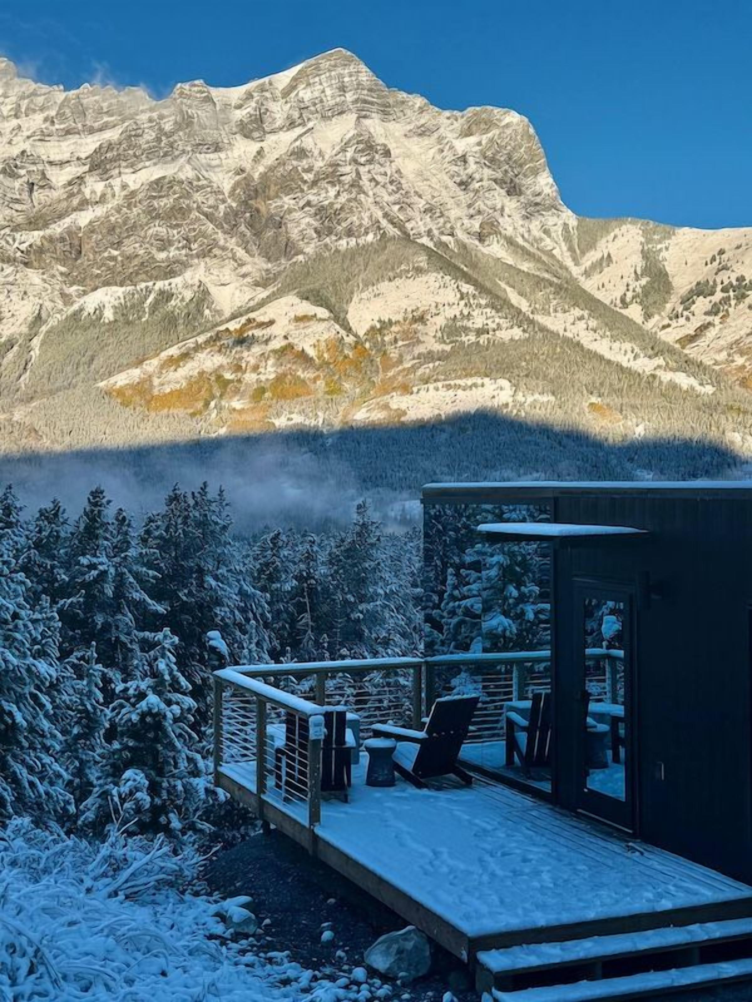 Varanda coberta por neve com uma cadeira e mesa, com vista para uma cordilheira.