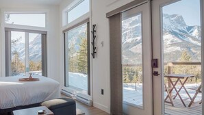 Ein Schlafzimmer mit einem großen Bett, einem Holznachttisch, einer Bank und Blick auf verschneite Berge durch große Fenster.