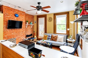 A compact living space with a brick wall, a ceiling fan, a television, a sofa with cushions, a small table, and a kitchen area with a sink and utensils.